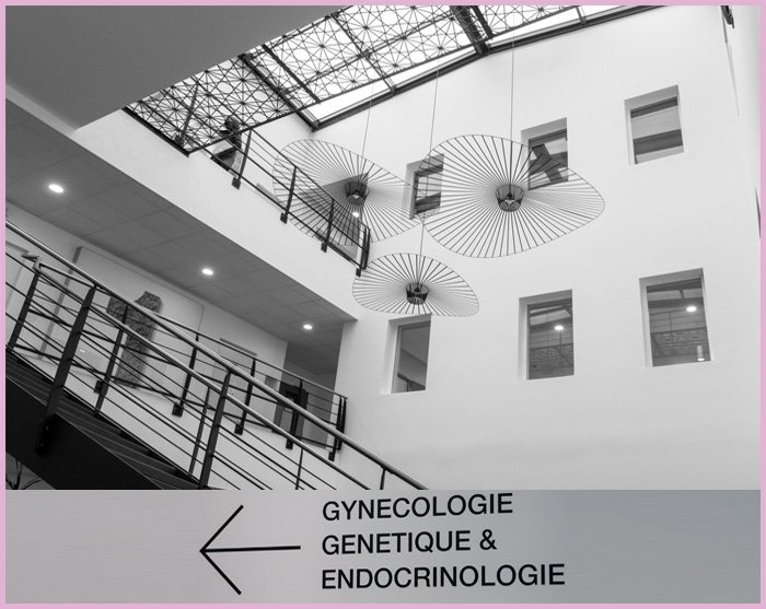 Le hall du cabinet : un atrium clair sous verrière, un escalier métallique, et un panneau indiquant la direction de la gynécologie.